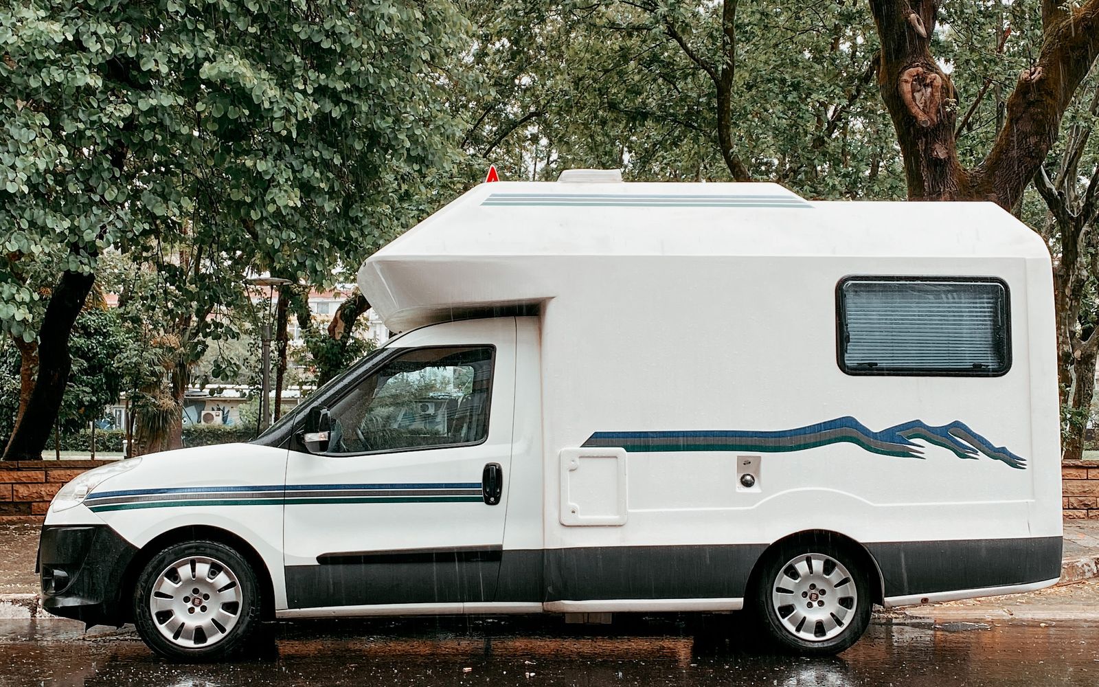 Ældre alkove-autocamper parkeret på asfalt ved høje træer